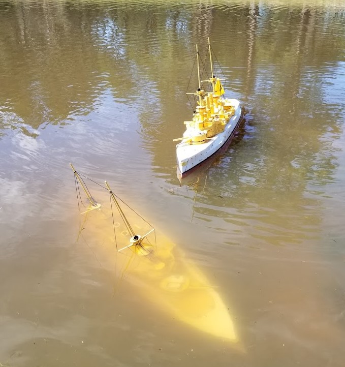 a sunk warship rests at the bottom of a shallow pond, with its masts still visible above the water