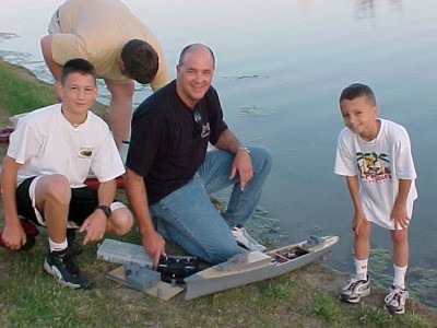 a man and two children pose with a model warship near a pond